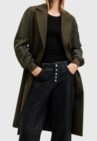 Gabardina verde oliva con un diseño estructurado, cuello oversized y mangas elásticas, combinada con pantalones negros de pierna ancha con botones plateados.