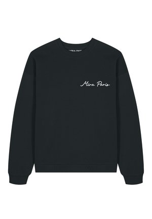 Jeune homme ajustant son col, portant un sweat noir avec le texte « Mira Paris » et un jean bleu clair sur fond blanc