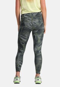 Donna che indossa leggings neri e verdi con un motivo astratto e sneakers con accenti neon, in piedi di spalle rispetto alla fotocamera.
