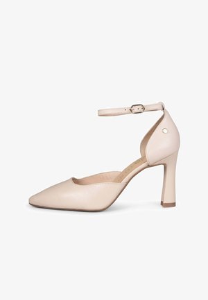 Zapato de tacón alto de cuero beige con punta en forma de pico, con correa en el tobillo y cierre de hebilla, mostrado desde el lado izquierdo sobre un fondo blanco.