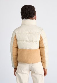 Pufferjack met een crèmekleurig bovenstuk en een tan, gestructureerde fleece middenstuk. Voorzien van een hoge kraag en elastische manchetten. Achteraanzicht wordt weergegeven.