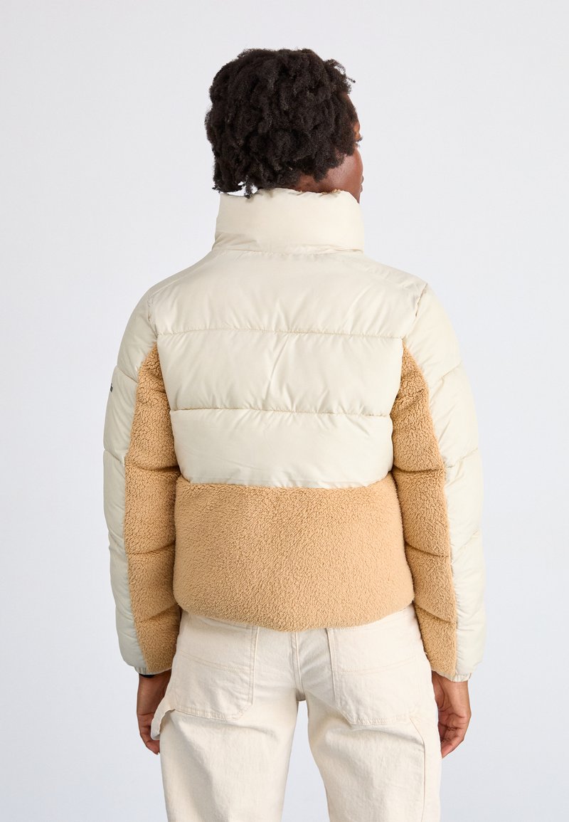 Kurtka puchowa z kremową górną częścią i jasnobrązowym, teksturowanym polarem w środkowej części. Oferuje wysoki kołnierz i elastyczne mankiety. Wyświetlany jest widok z tyłu.