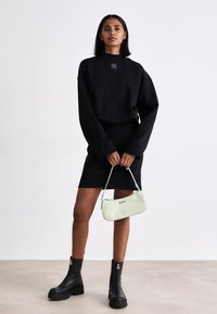 Čierny oversized sveter a priliehavá sukňa doplnená o robustné čierne čižmy. Zelená kabelka s detailom loga. Jednoduchý, moderný dizajn.