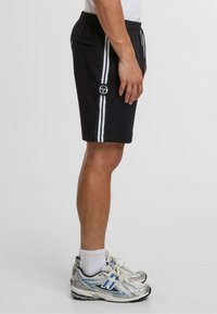 Černé sportovní šortky s bílými pruhy, elastickým pasem, bočními kapsami a logem na levém předním dílu. Kombinováno se stříbrnými a modrými teniskami.