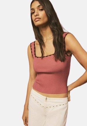Femme aux longs cheveux foncés portant un haut rose côtelé sans manches avec bordure noire et un pantalon blanc avec détails cloutés, posant avec une main sur la hanche.
