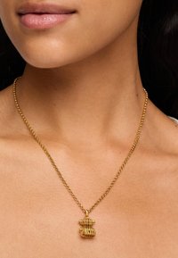 Złota naszyjnik z teksturowanym wisiorkiem o falistym wzorze, zawieszony na delikatnym łańcuszku. Zbliżenie na szyję noszącej.