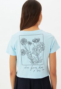 Camiseta corta azul claro de algodón, con un dibujo en línea negro de flores y el texto "donde florecen las flores, también florece la esperanza."