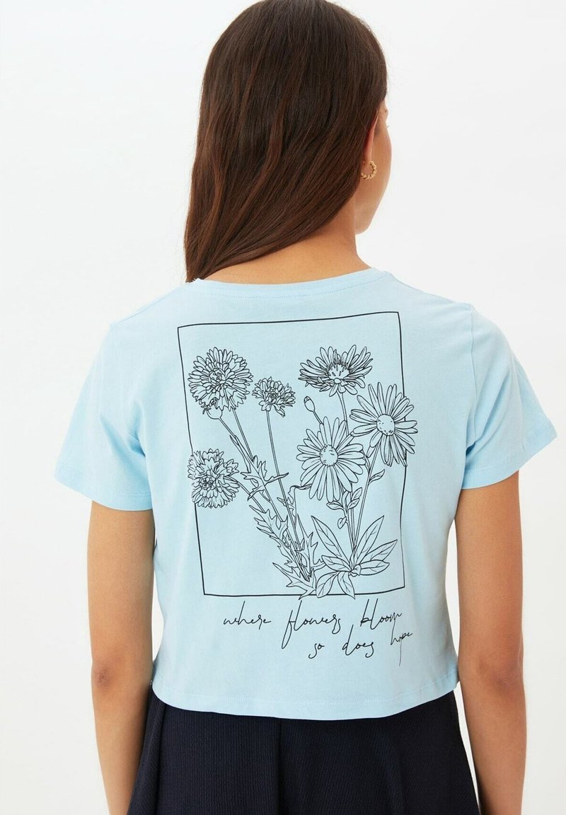 Camiseta corta azul claro de algodón, con un dibujo en línea negro de flores y el texto "donde florecen las flores, también florece la esperanza."
