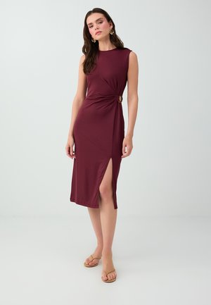 Femme debout portant une robe midi bordeaux sans manches avec une fente latérale et un détail boucle dorée, associée à des sandales beiges et des boucles d'oreilles créoles dorées.