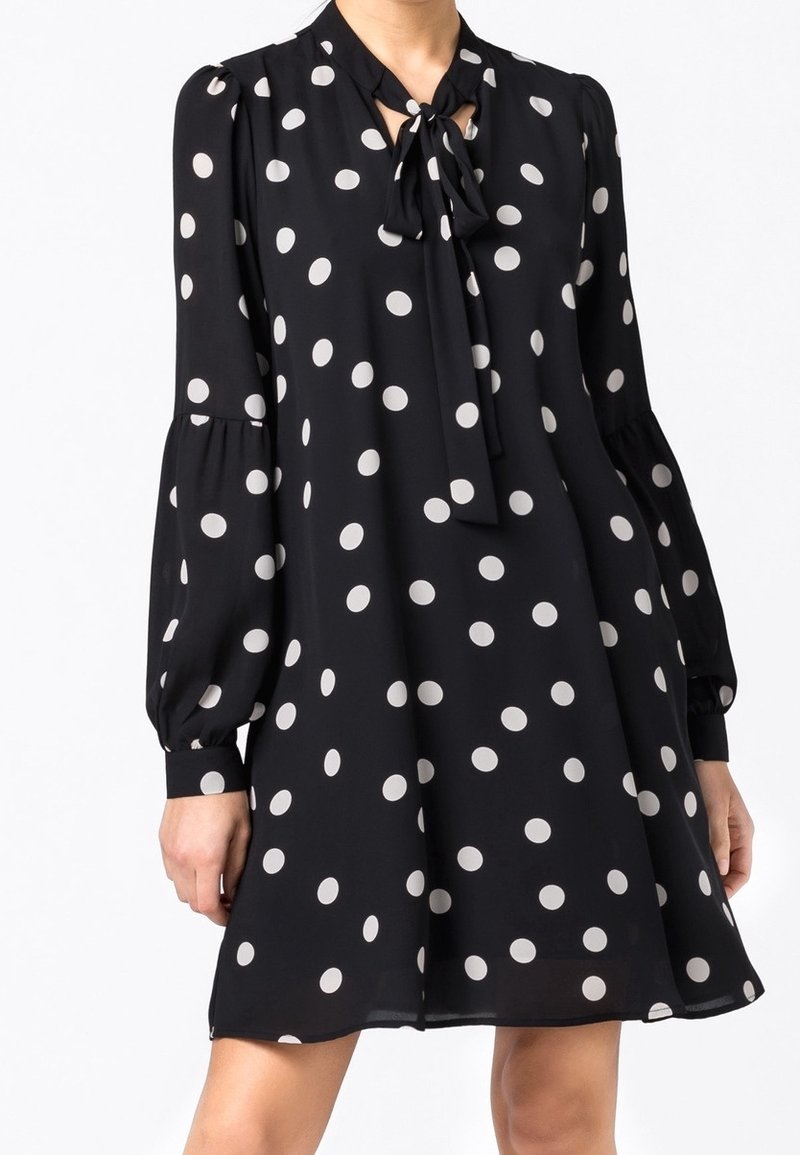 Femme portant une robe noire à manches longues avec des pois blancs et un nœud noué au niveau de l'encolure.