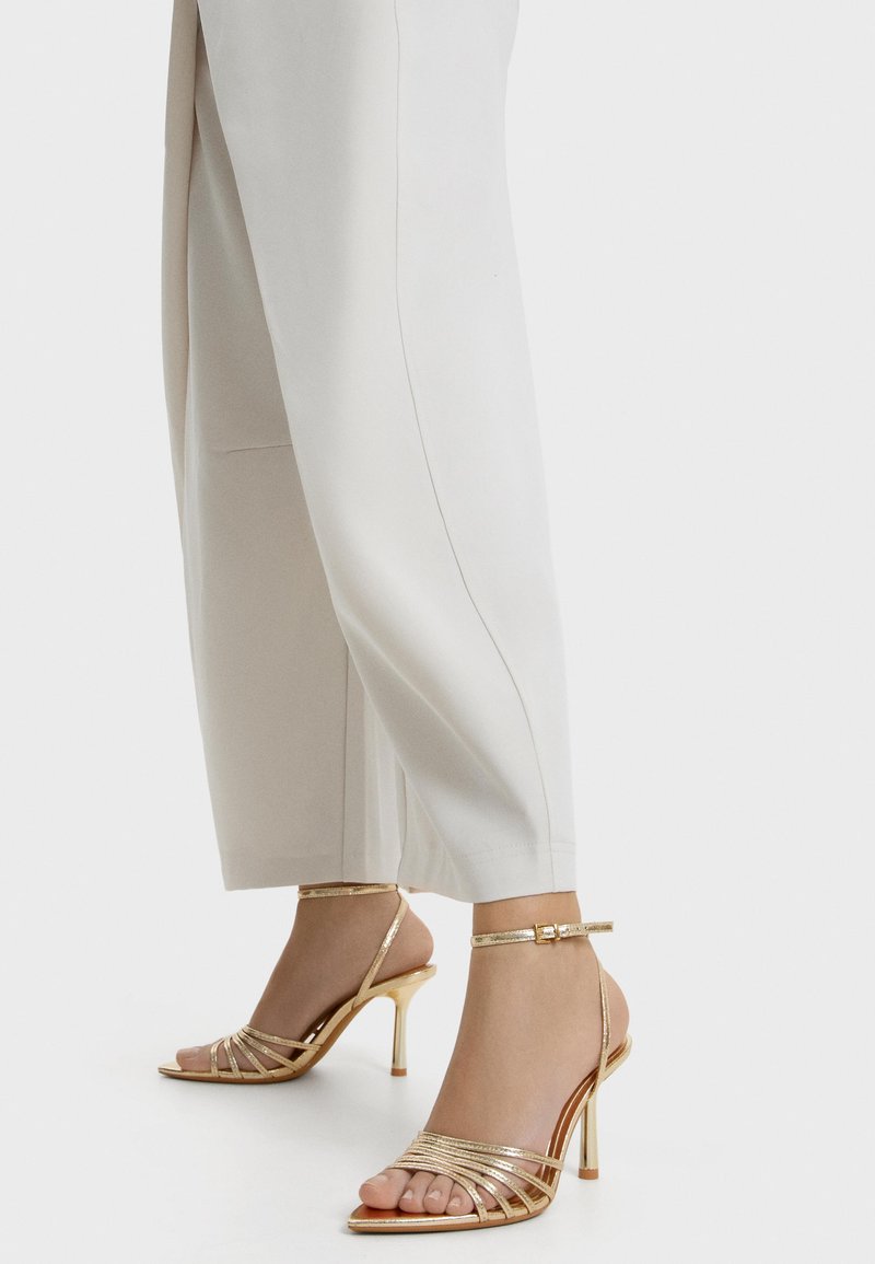 Picioare purtând sandale aurii cu toc înalt și barete, asortate cu pantaloni albi largi, pe un fundal discret deschis.