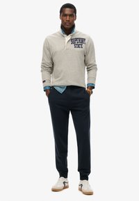 Suéter gris con un logo en navy "SUPERDRY STATE", superpuesto a una camisa de chambray azul claro, combinado con joggers oscuros en navy y zapatillas blancas.