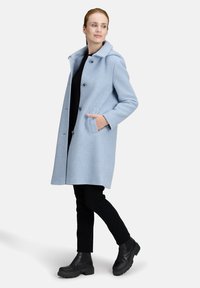 Cappotto di lana azzurro chiaro con colletto, chiusura a bottone e due tasche laterali. I pantaloni neri aderenti e gli stivali neri gommoni completano il look.