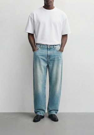 Άνδρας που φοράει απλό λευκό κοντομάνικο T-shirt, ανοιχτό μπλε ξεβαμμένο φαρδύ τζιν, μαύρα μοκασίνια, στέκεται με τα χέρια στις τσέπες σε απλό φόντο.