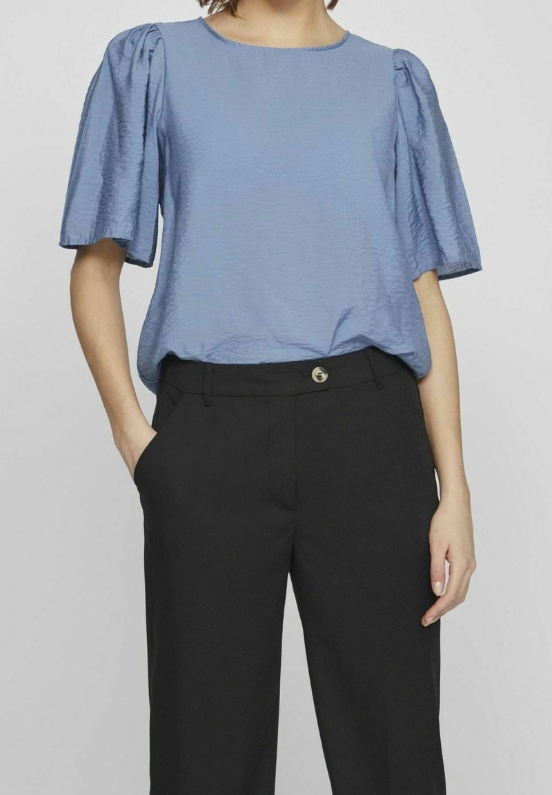 Haut bleu clair à manches courtes avec une finition texturée, encolure ronde et manches bouffantes, associé à un pantalon noir avec un détail de bouton.