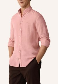 Homme portant une chemise en lin rose pâle à boutons avec les manches retroussées et un pantalon marron foncé, mains dans les poches, sur un fond neutre.