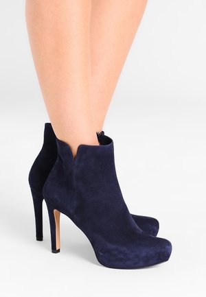 Woman wearing navy blue suede high-heeled ankle boots with pointed toes and a V-cut front opening.