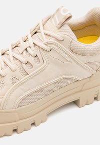 Zapatilla deportiva beige que presenta una combinación de suave ante y malla, suela de goma gruesa con un agarre texturizado y un logotipo destacado de la marca en la lengüeta.