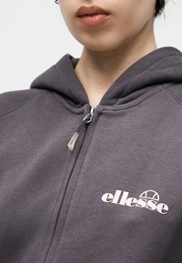 Κοντινή λήψη ενός ατόμου που φοράει σκούρα γκρι φούτερ με φερμουάρ και λευκό λογότυπο «ellesse» στο στήθος.