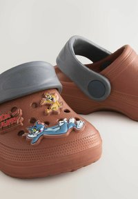 Sabots de style Crocs bruns avec une sangle grise, ornés de charms représentant les personnages Tom et Jerry, avec des trous de ventilation et une surface texturée.