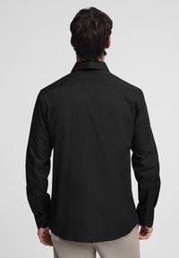 Maglietta nera a maniche lunghe con una texture liscia, collo classico, polsini con bottone e dettagli di cucitura precisi, vista dal retro.