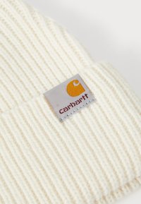 Krema košara iz pletiva, izrađena od teksturirane pletene tkanine, sa kvadratnom Carhartt oznakom koja ima narančasti logotip i crveni tekst na sivoj pozadini.