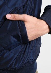 Marinblå isolerad jacka med kviltat mönster, sidofickor, ribbade ärmar och en slät textur. Hand delvis inskjuten i fickan.