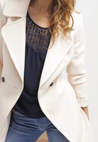 Cappotto sagomato rosa pallido con colletto a revers, bottoni doppi e un top blu navy con pizzo sottostante. Indossa jeans blu medio.