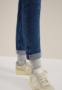 Jeans de mezclilla azul oscuro con los puños enrollados que revelan calcetines grises, combinados con deportivas beige claro con acentos blancos y un logo.