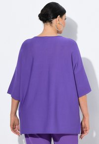 Lila übergroße Bluse mit kurzen Ärmeln, glatter Textur und seitlichen Schlitzen. Zubehör besteht aus großen Creolen. Modell von hinten angesehen.