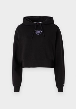 Svart cropped hettejakke med snorhette. Har et lilla brodert hjerte-logo på brystet og ribbestrikkede mansjetter. Myk stofftekstur.