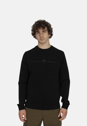 Jonge man met krullend haar in een zwart North Sails sweatshirt en olijfgroene broek, staande tegen een effen lichte achtergrond.