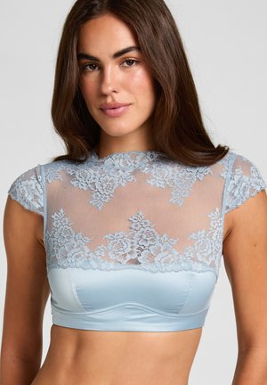 Jonge vrouw met lang bruin haar, die een lichtblauw kanten crop top draagt met bloemenpatronen en korte mouwen tegen een effen achtergrond.