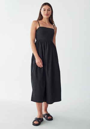Musta olkaimeton mekko, jossa on tiukka bodice ja fruntersi vyötärö, ulottuen puoleen vasikkaan. Yhdistetty mustien nauhakenkien kanssa.