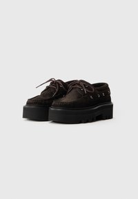 Scarpe in suede marrone con una robusta suola piatta nera. Presentano lacci rotondi, occhielli in metallo e un design cucito in stile mocassino.