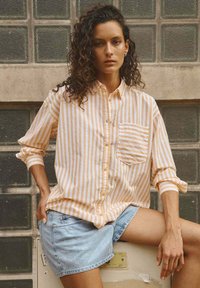 Jeune personne avec des cheveux bouclés portant une chemise à rayures jaunes et blanches à boutons et un short en denim, assise de manière décontractée contre un mur avec des blocs de verre.