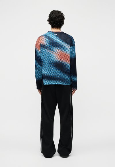 Homme vu de dos portant un haut à manches longues avec des points abstraits bleus et orange, et un pantalon noir avec des bandes blanches sur les côtés.