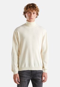Pull à col roulé crème en tissu tricoté texturé, avec des manches longues et une coupe décontractée. Porté avec un jean gris foncé.