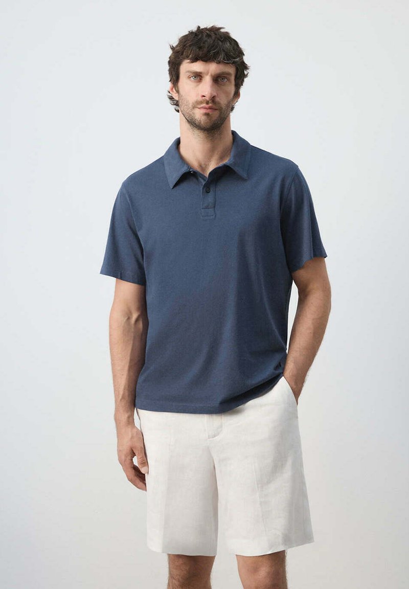 Homme aux cheveux courts et foncés, portant un polo bleu marine et un short blanc, debout avec une main dans la poche devant un fond uni.