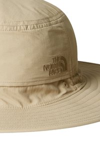 Sombrero tipo bucket beige hecho de tejido ligero, con un ala ancha, detalles de costura y un logo en relieve en la parte frontal.