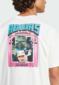 White cotton t-shirt featuring a large, colorful graphic of a green character riding a duck, framed by pink with pixelated accents and text.