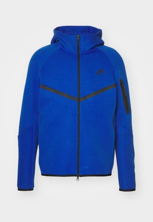 Blaues Kapuzenjacke mit Reißverschluss, das einen hohen Kragen, schwarze Akzente und eine Reißverschlusstasche am linken Arm hat. Hergestellt aus einem weichen Stoffgemisch.