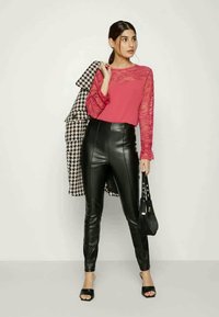 Top de renda rosa de mangas longas combinado com calças pretas de couro sintético de cintura alta, segurando uma bolsa preta, com um casaco em padrão pied-de-poule a escorregar pelo ombro.