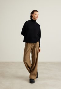 Schwarzer Strickpullover mit Rollkragen, kombiniert mit hochtaillierten, weiten Beigetönen und schwarzen Plateau-Schuhen, vor einem neutralen Hintergrund.