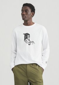 Λευκό μακρυμάνικο μπλουζάκι με μαύρο γραφικό κρανίο με κουκούλα και πλαϊνή κορδέλα «VO.COM», φορεμένο με παντελόνι ελιάς πράσινου χρώματος.