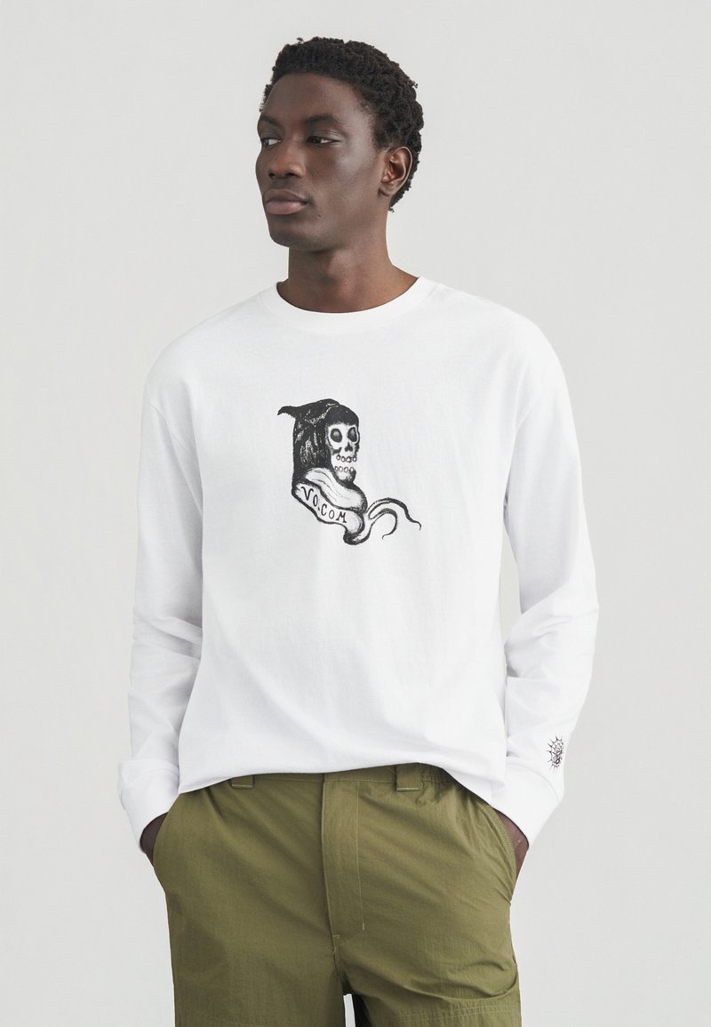 Λευκό μακρυμάνικο μπλουζάκι με μαύρο γραφικό κρανίο με κουκούλα και πλαϊνή κορδέλα «VO.COM», φορεμένο με παντελόνι ελιάς πράσινου χρώματος.