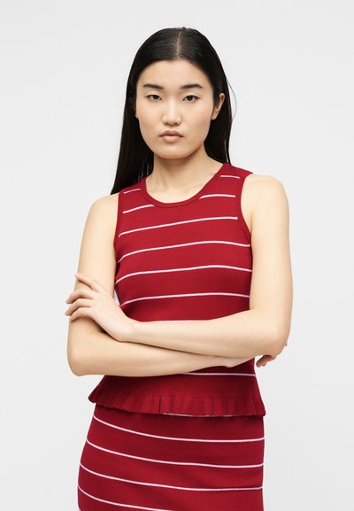 Νέα γυναίκα με μακριά μαύρα μαλλιά που φοράει ένα κόκκινο πλεκτό τοπ χωρίς μανίκια και φούστα με λεπτές λευκές οριζόντιες ρίγες, με ταυτισμένα χέρια.