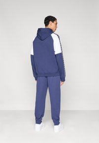 Σετ αθλητικής φόρμας σε σκούρο μπλε, που περιλαμβάνει φούτερ με κουκούλα και αντίθετες λευκές πινελιές, παντελόνι που ταιριάζει και λευκά αθλητικά παπούτσια.