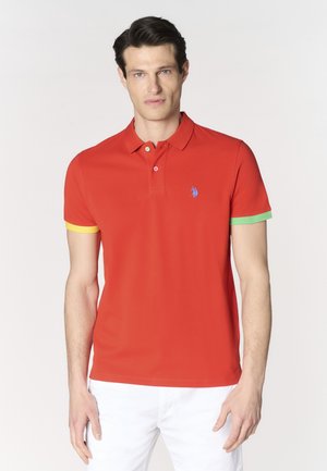 Uomo che indossa una polo rossa con polsini gialli e verdi, logo ricamato blu sul petto e pantaloni bianchi, in piedi su sfondo bianco.