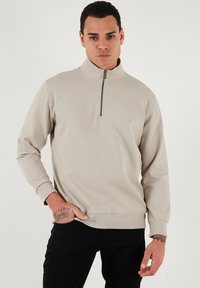 Svetlo béžový pulóver s kvartovým zipsom vyrobený z textúrovaného materiálu s dlhými rukávmi a kovovým zipsom. Obsahuje rebrované manžety a voľný strih.